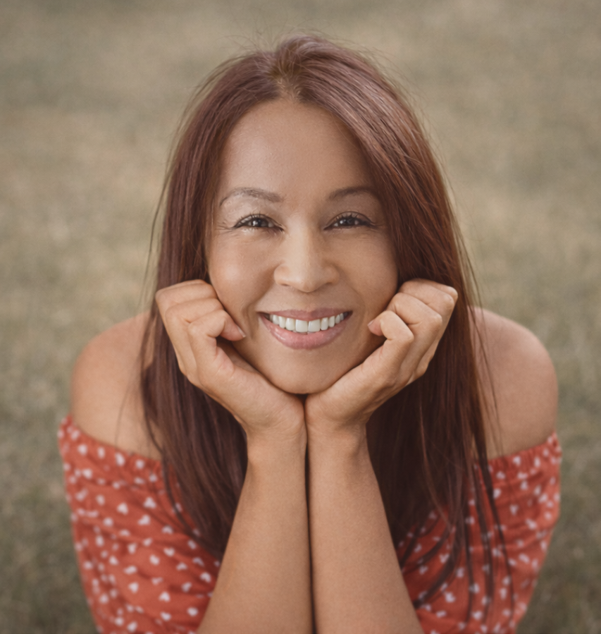 A woman smiles brightly at the camera while resting her chin in her hands.